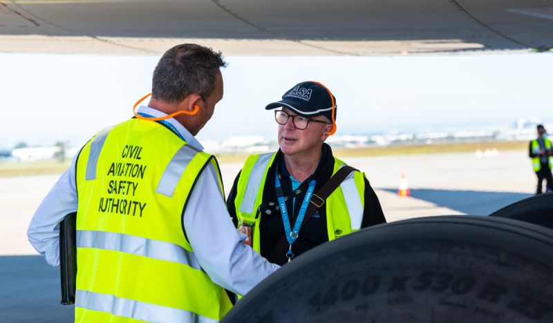 Two individuals in high-visibility vests on an airport tarmac with a large aircraft tire in the foreground.
